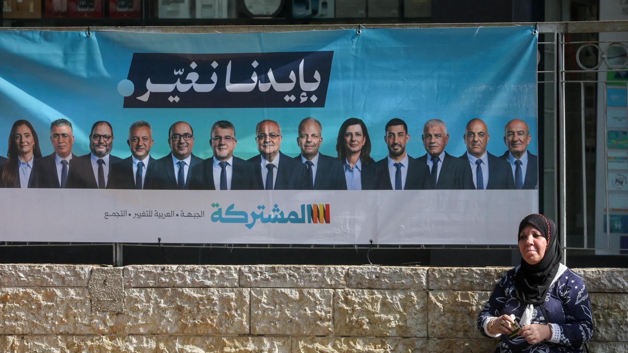 A woman sits near an election campaign banner featuring leaders of the Joint List, in Nazareth, Israel (10 October 2026)