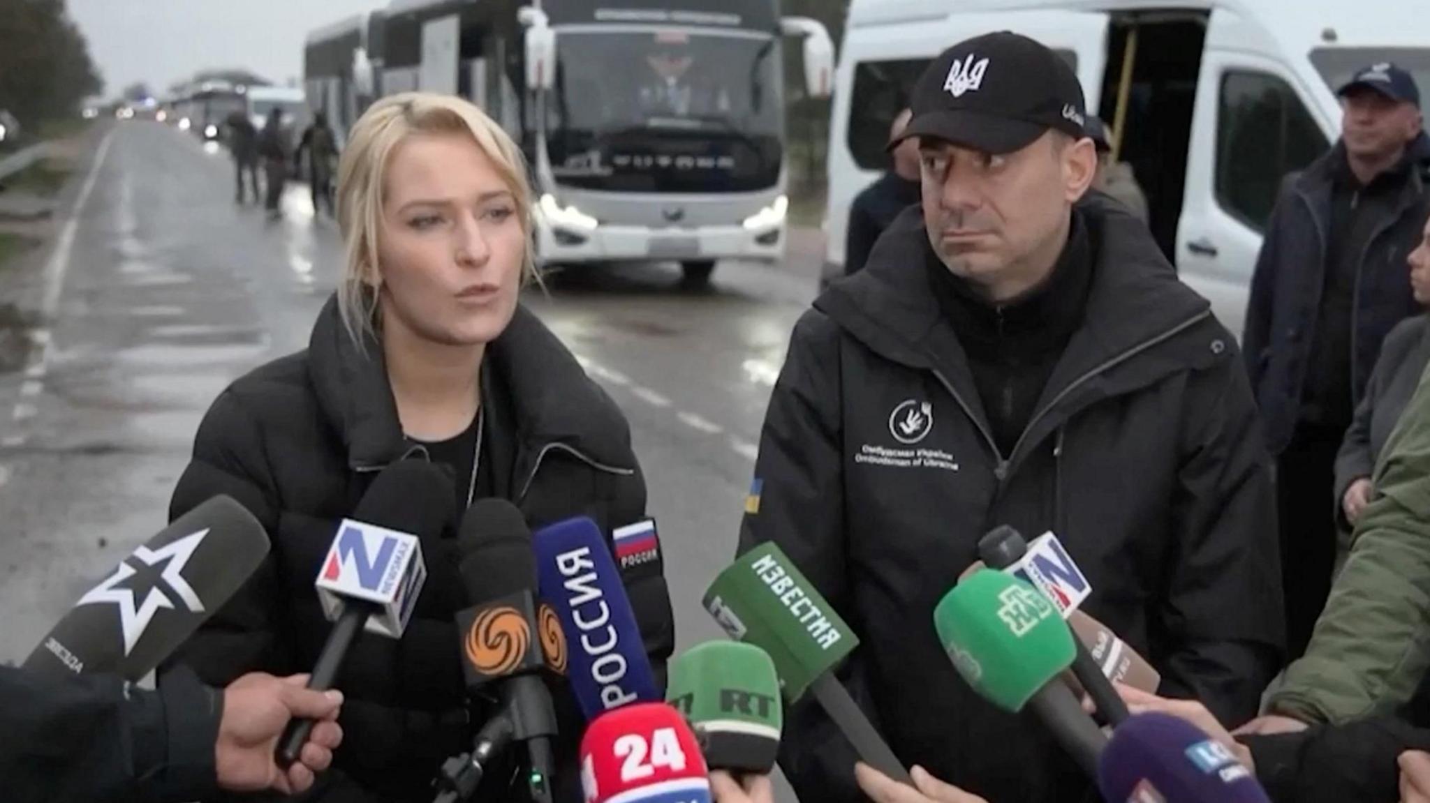 Yana Lantratova is on the left, standing next to Dmytro Lubinets on the right. There are several microphones with the logos of mainly Russian TV channel pointing at them. Lantratova has blonde hair pulled back into a ponytail, and wears a black jacket with the flag of Russia on one arm. Lubinets wears a black jacket and a black baseball cap with a Ukrainian national emblem on it.