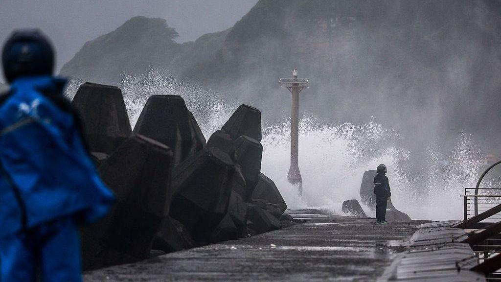Large waves crash against a coastal breakwater during stormy weather, sending spray high into the air. Massive concrete tetrapods line the left side of the seawall, while two people wearing blue rain gear stand on the wet waterfront.