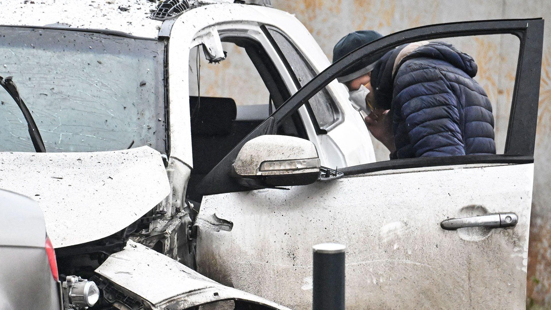An investigator wearing a face mask and a blue padded jacket looks into a destroyed car in southern Moscow on December 22, 2025. The car is white and has a shattered windscreen and damage to the roof and bonnet.