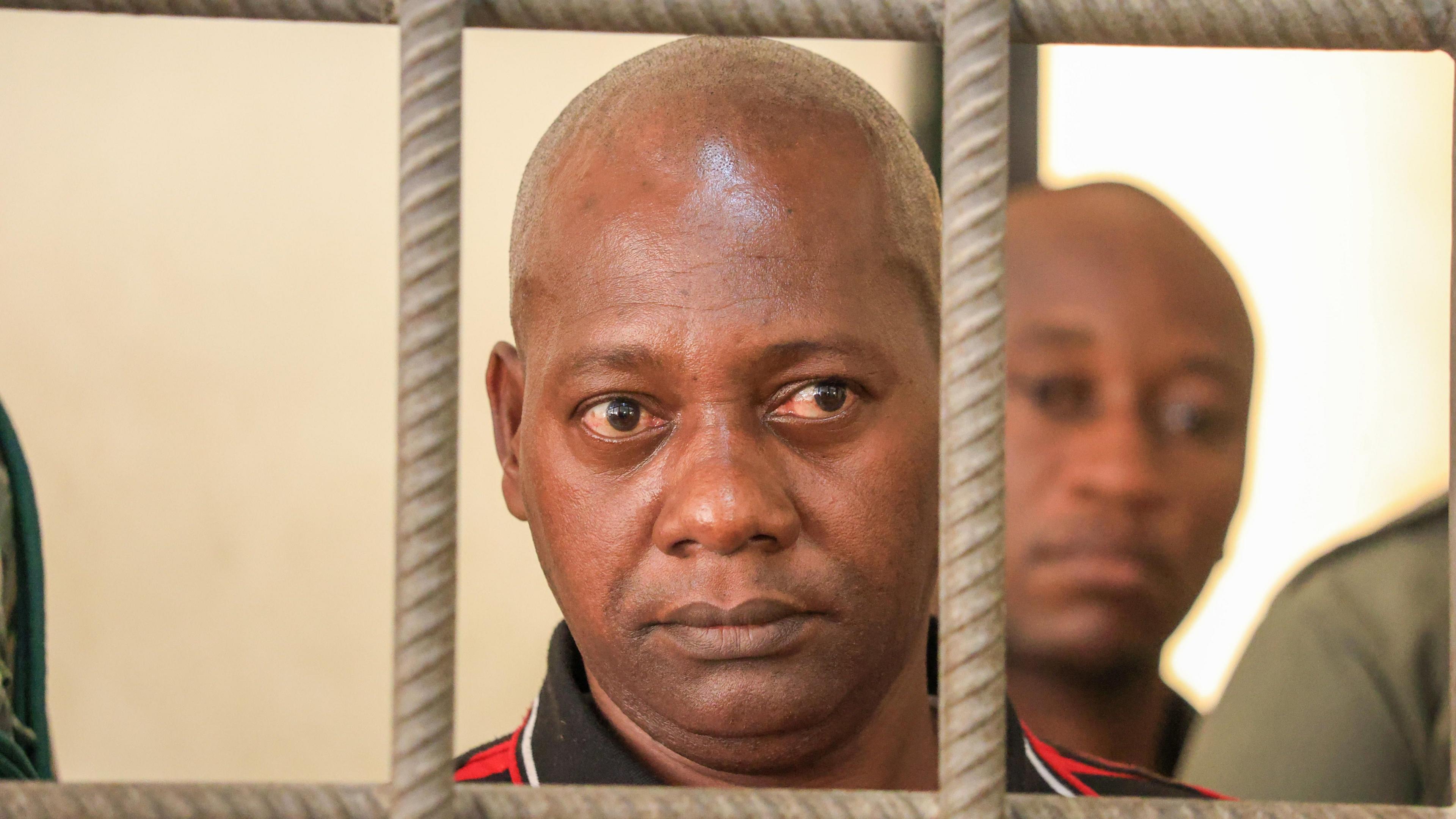 A close shot of a bald man looking through the bars of a cell