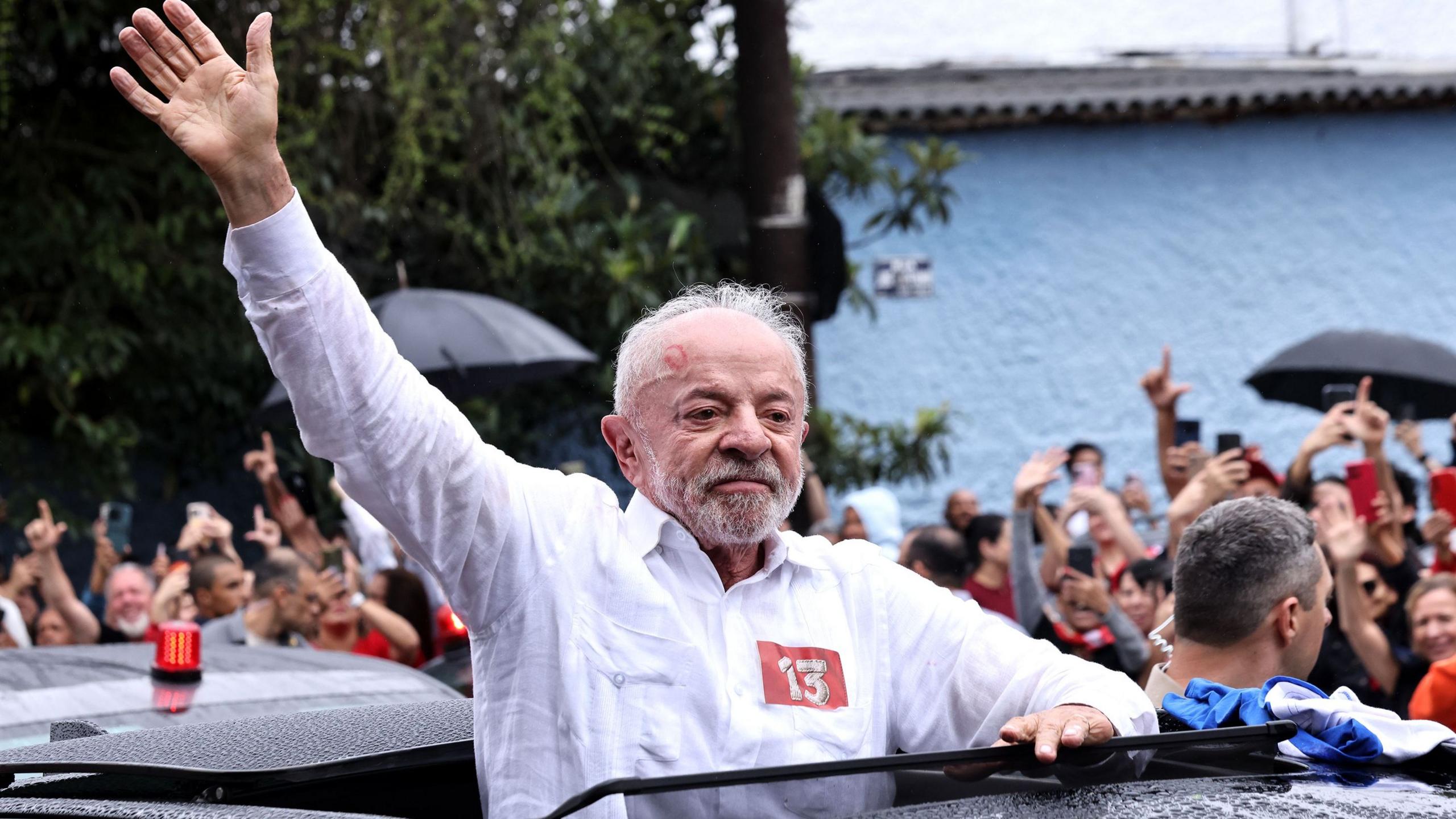 Lula is pictured with the upper part of his body from the roof of his car, wearing a white shirt extends his right arm to greet hundreds of supporters