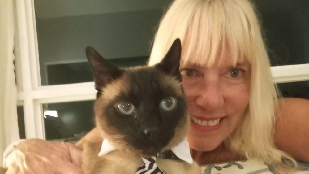 A Siamese cat wearing a black-and-white striped necktie and white collar sits in the foreground while being held indoors by a woman.