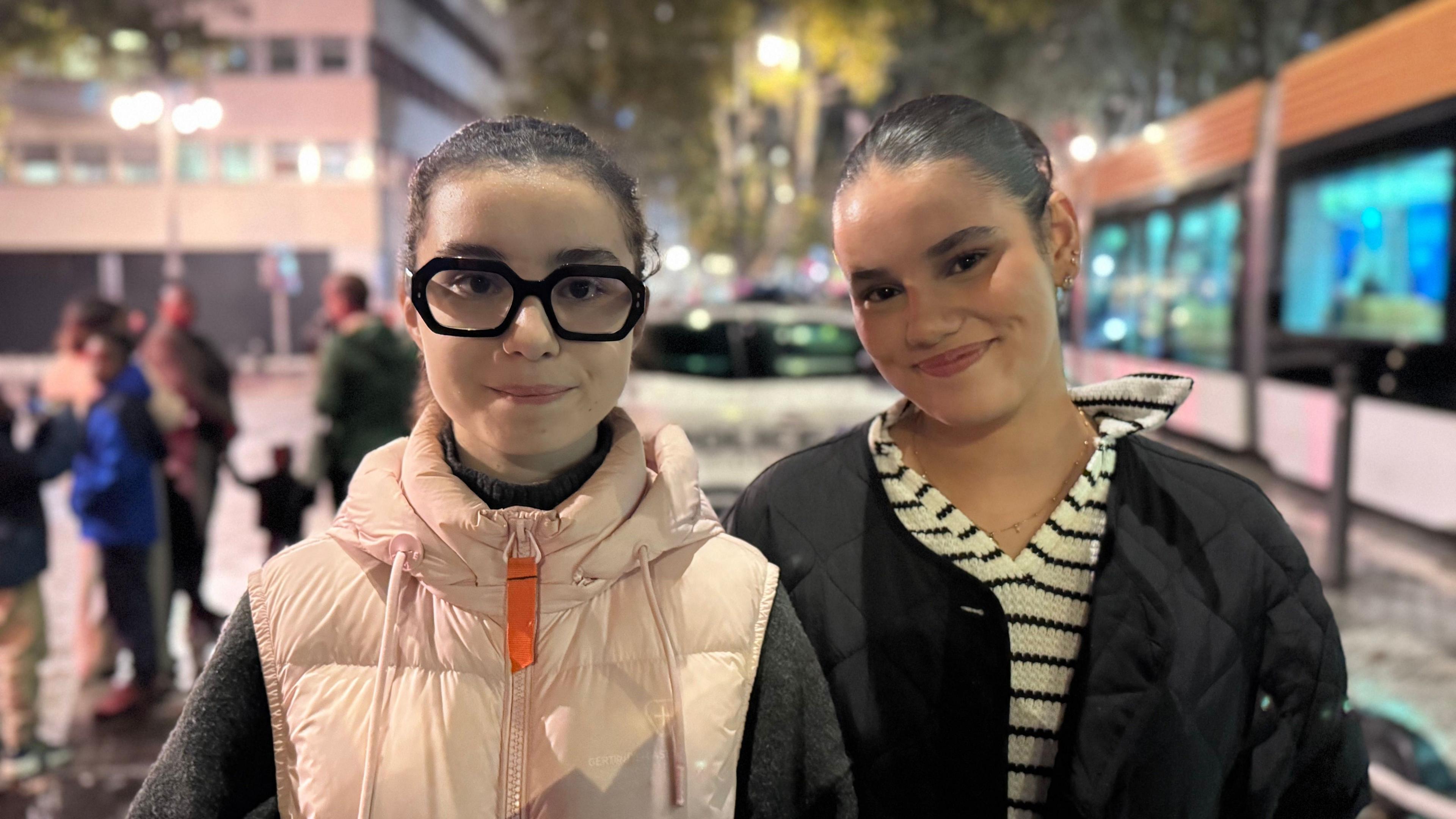 Two young women pictured in Marseilles, smiling at the camera in front of a tram stop at night. Maryam is on the right. She has dark brown hair which is pulled back, and wears a black jacket above a v-neck striped black and white top. Jade is on the left. She also has dark hair pulled back, and wears thick black-framed glasses. She is wearing a light pink gilet above a black jumper.