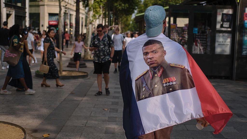 man walks through Paris with flag on back showing Mbappe in military uniform