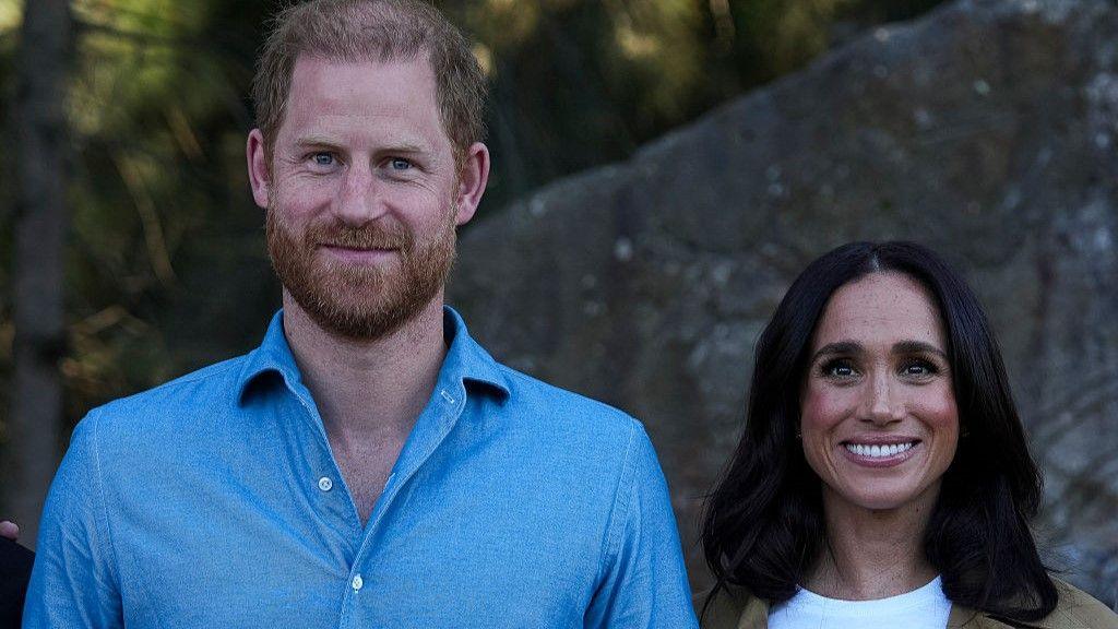 Harry stands next to Meghan. He is wearing a light blue shirt. They are both smiling.