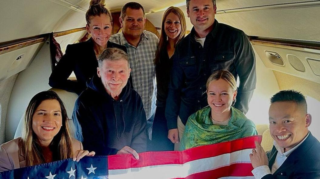Stephen Hubbard (second left, sitting), pictured with members of his family holding a US flag inside a plane. There are eight people in the photo, four standing and four sitting.
