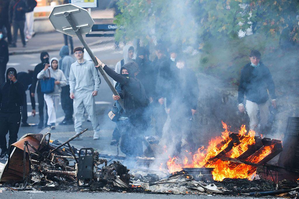 A road sign is hurled into burning wreckage in the street by a masked and hooded man