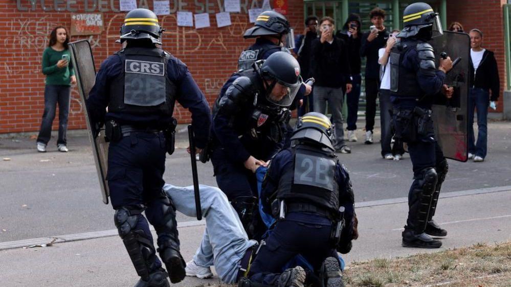A protester is seen bundled to the ground with two riot police holding him as students in the background film on their mobile phones