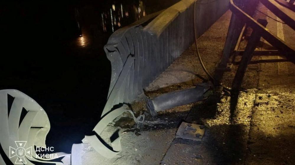 An image of damage caused to the railings of the Southern Bridge in Kyiv. The metal is bent and ripped and there is debris on the floor. It is dark outside.