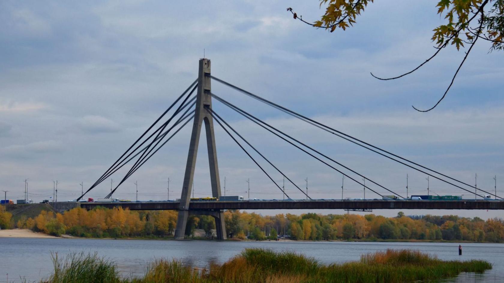 A view of the Pivnichnyi Bridge in Kyiv, Ukraine. There are cars driving across it and water running below with orange and green trees lining the banks.