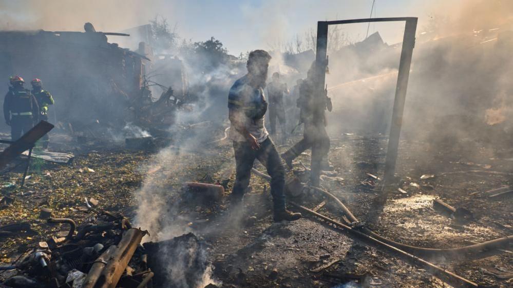 Several emergency workers can be seen standing amidst rubble and smoke, in north-eastern Kharkiv following a strike. There is smoke in the air and damaged metal all over the ground.