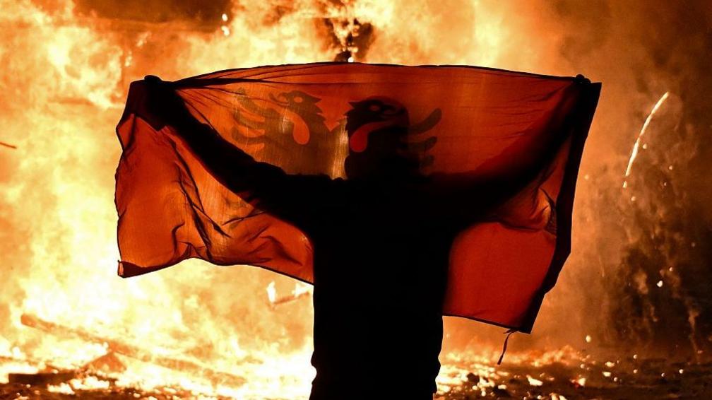A protester holds an Albanian National flag in front of a burning kiosk during an anti-government demonstration in central Tirana on October 15, 2026