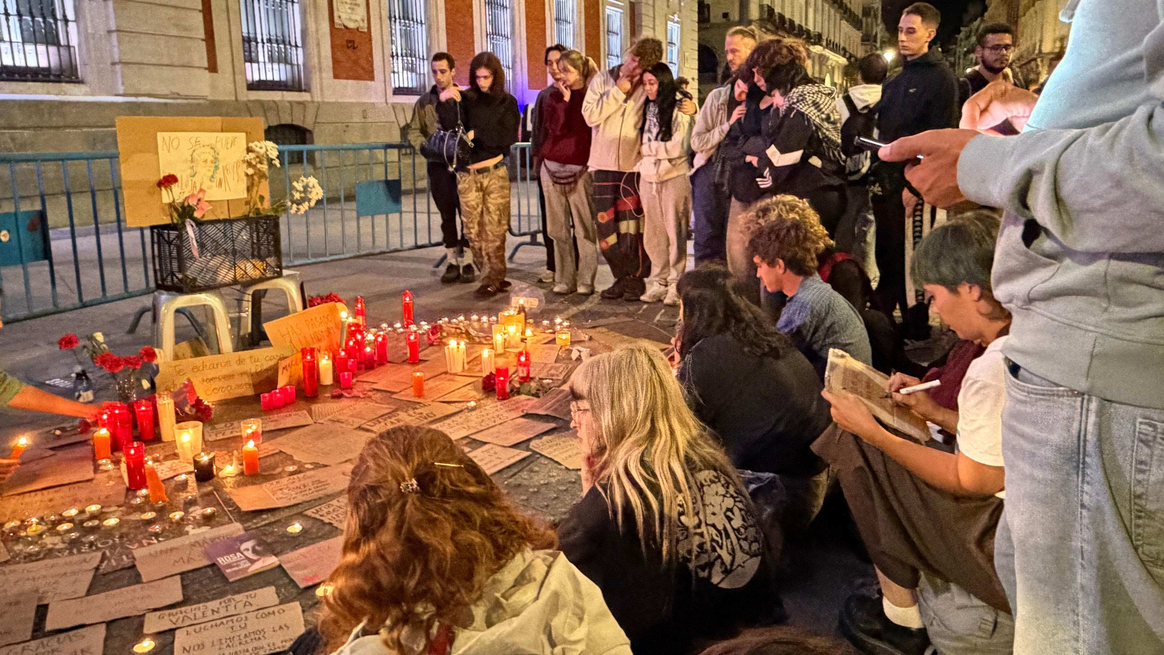 People gather around a vigil where red candles and written tributes sit on pavement near a fence.