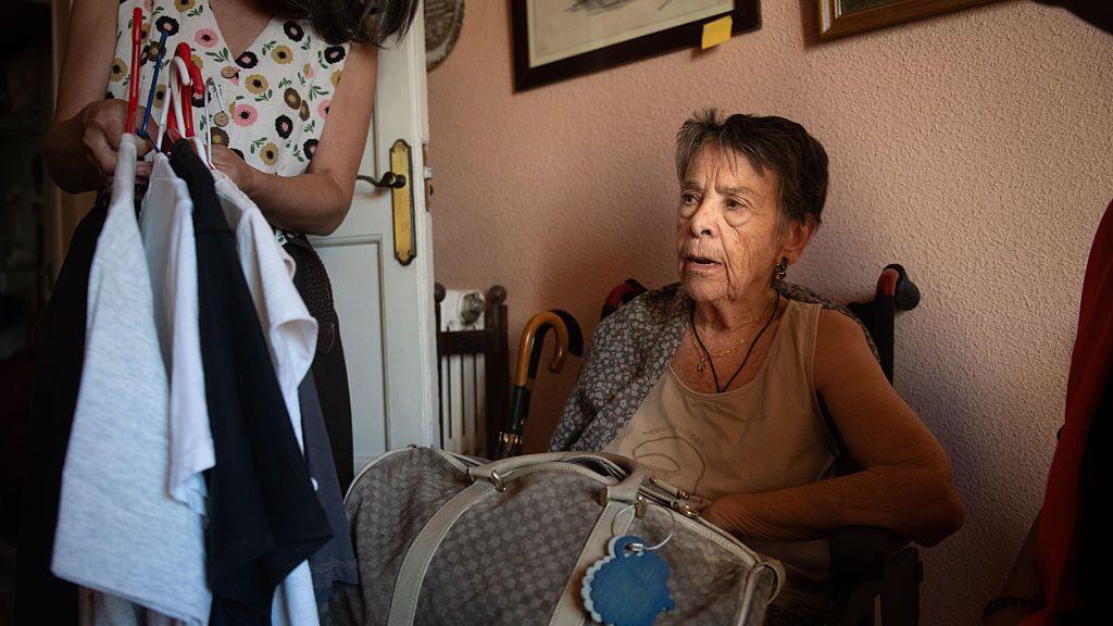 Maricarmen Abascal sits in a chair in her flat ahead of her eviction, in Madrid, Spain on 23 September. She has short, dark hair and is wearing a cream vest. A holdall bag is on her lap. A woman whose face is out of shot is holding up clothes on a hanger next to Abascal.