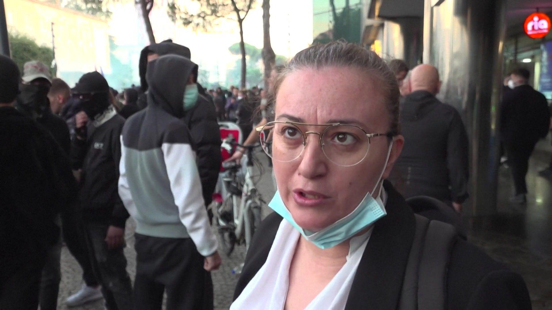 Enida Bimi speaking to a camera, with a mask below her chin, in front of various protesters