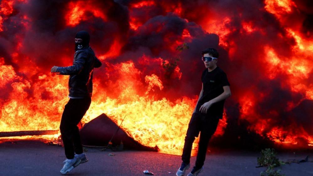 Two young men, one wearing a balaclava and another in big sunglasses, stand in front of a huge plume of fire