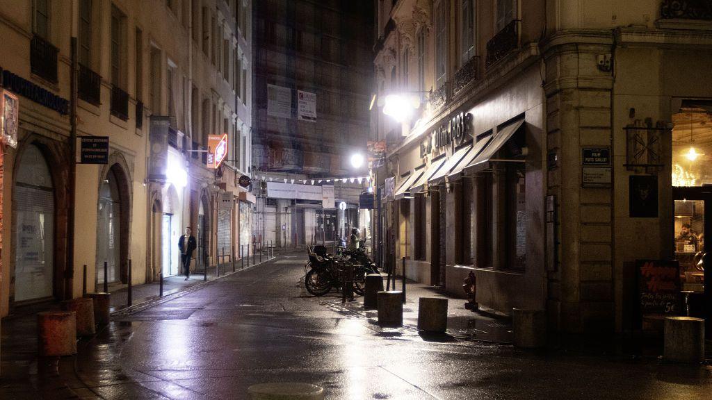A man walks down a street in Lyon at night, where motorbikes are parked in front of a shop.