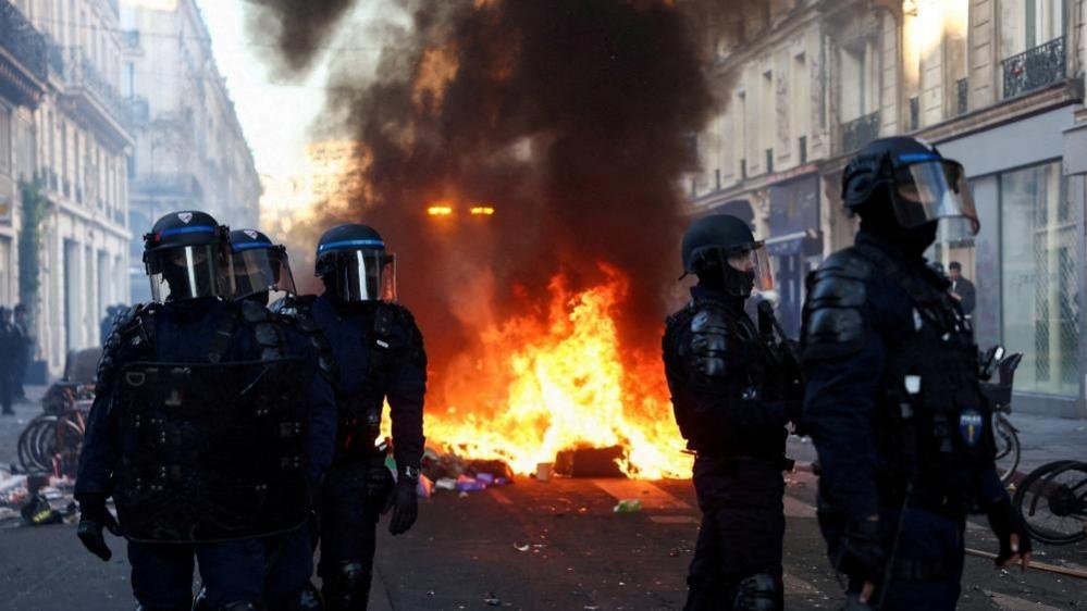 A fire burns as police officers in riot gear stand guard in Paris