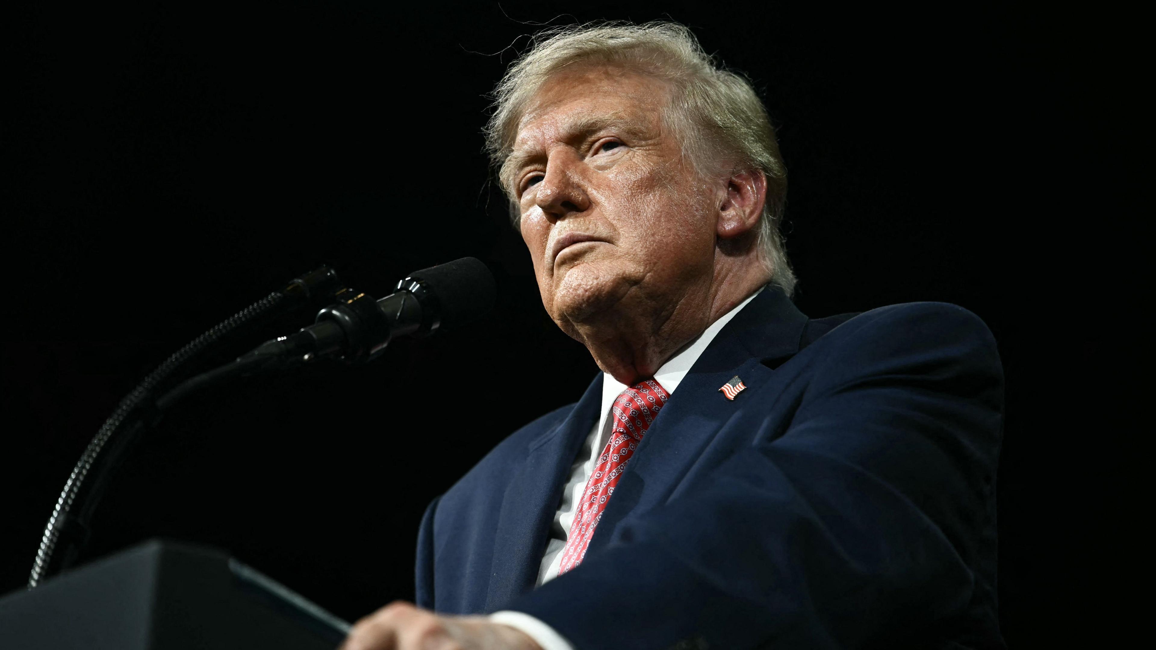 President Trump stands against a black background behing a black podium/ He is wearing a white shirt, blue suit coat with an American flag pin and a red print tie. He is not smiling and is gripping the edges of the podium. He is photographed from below the podium looking up.