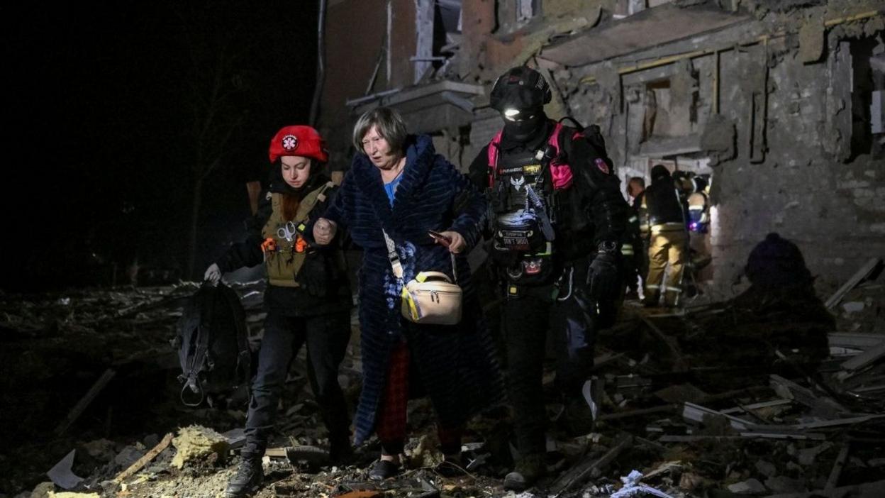 Wide shot showing a middle-aged woman walking away from a residential building that has been damaged by Russian strikes at night. She is holding the hands of two rescue workers who are helping her walk over the rubble.
