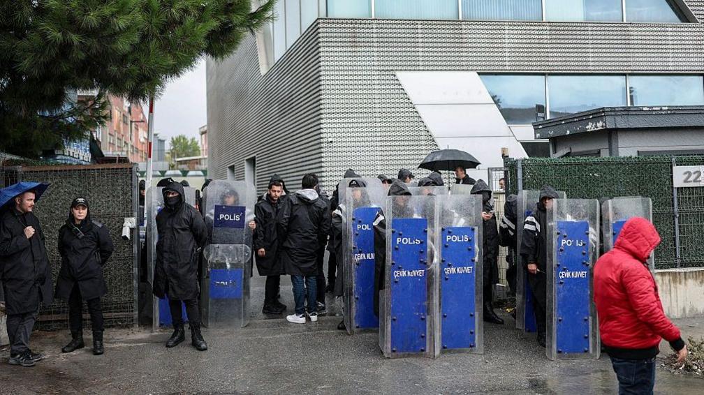 A row of riot police stood outside in the rain outside a grey office building.
