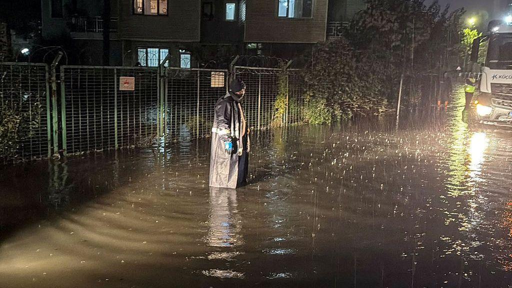 A woman in rain gear stands in flood water at night. The water is up to her calves