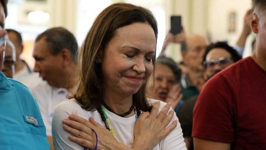 A woman in white crosses her arms on her chest with her hands pointed towards her shoulders and her eyes closed