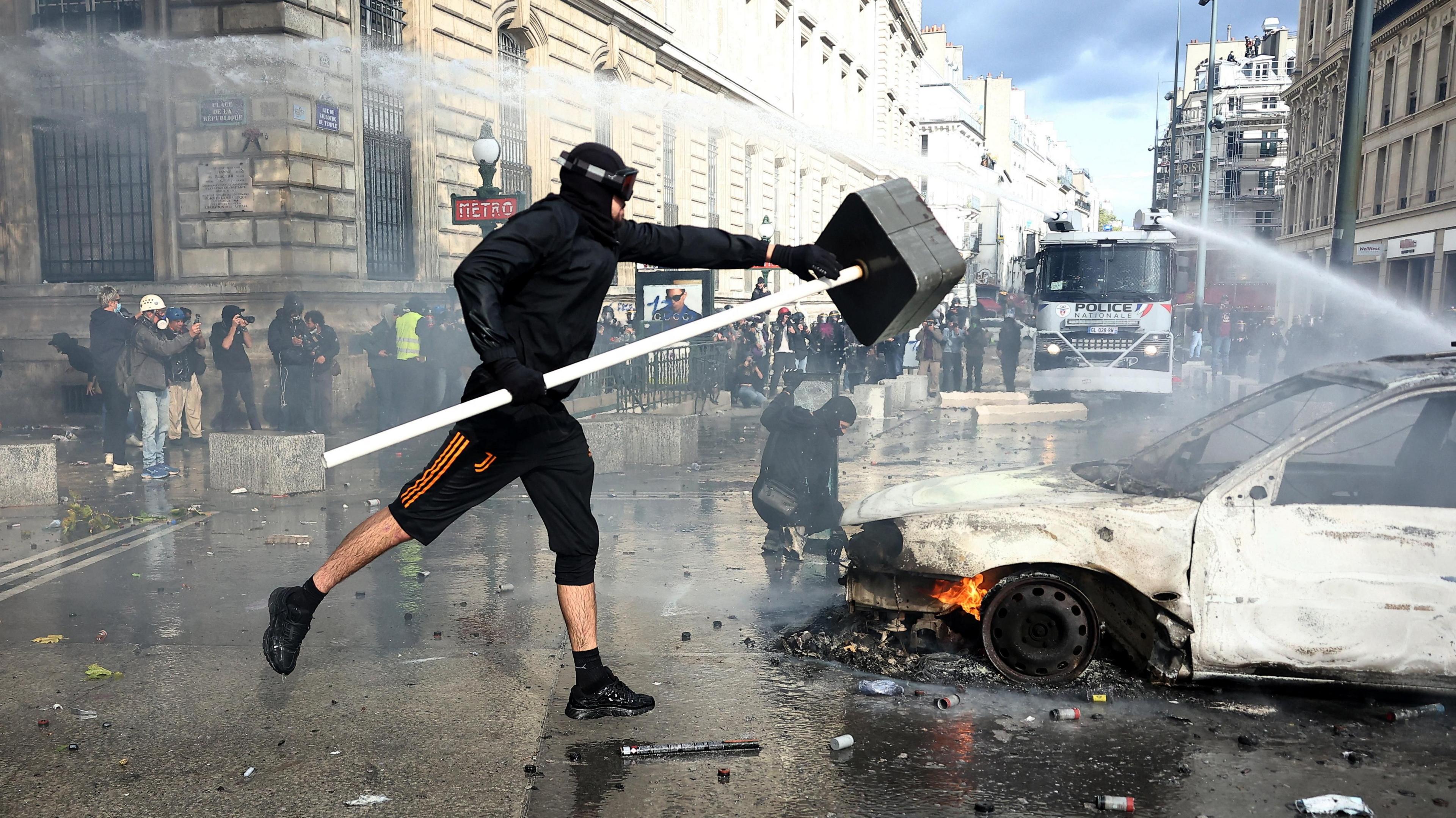 A masked protester throws an object onto a burning car as French riot police use a water canon amid clashes during a demonstration at the Place de la Republique in Paris