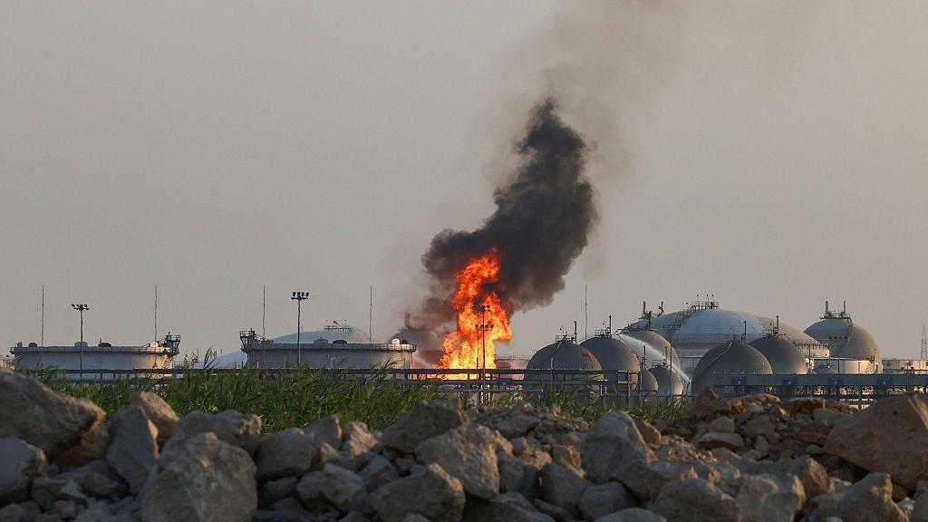 Flames and thick black smoke rise from an Aramco oil facility near Riyadh, with storage tanks visible behind a rocky foreground