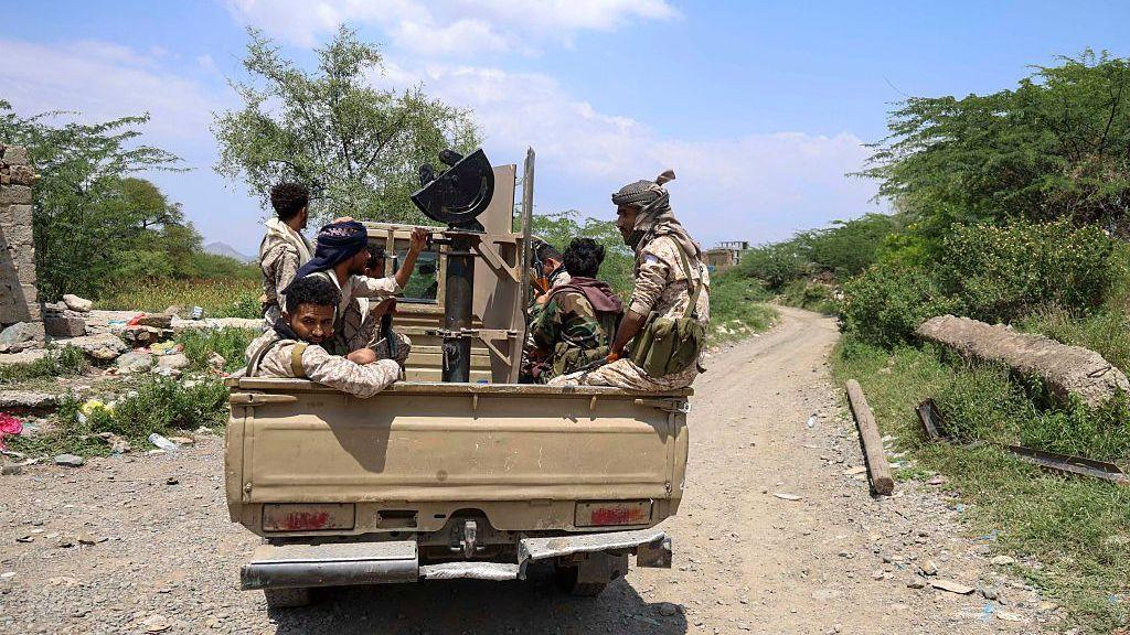 Several people ride in the back of a pick-up truck carrying mounted equipment along an unpaved road lined with trees and scrub