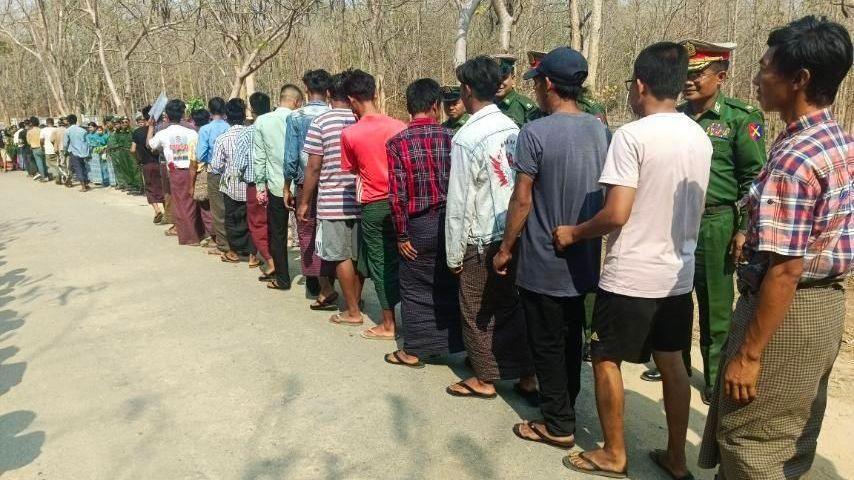 A long line of men standing on the side of a road, with soldiers in uniform watching over them