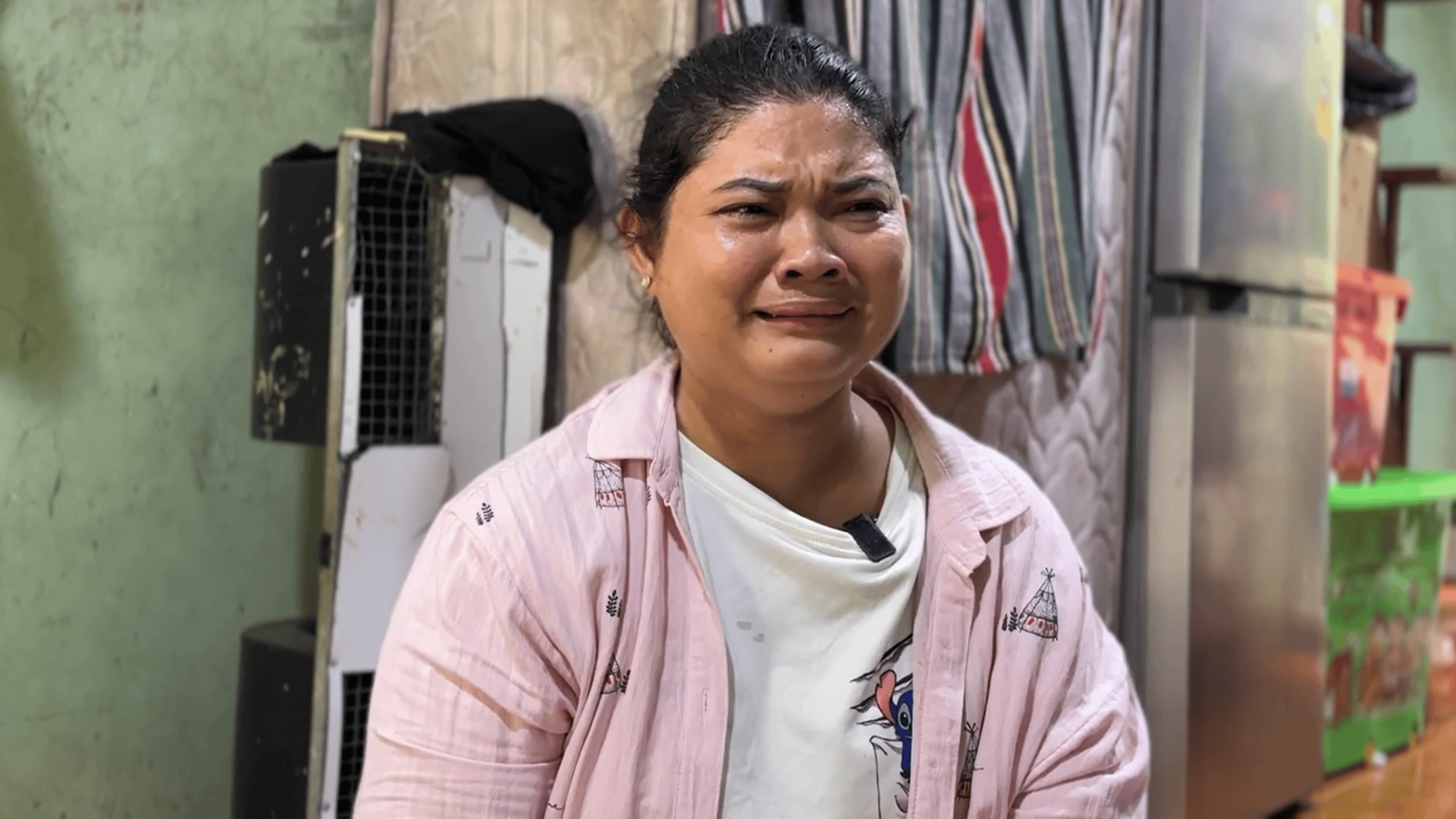 A woman wearing a white t-shirt under a pink overshirt grimaces in pain. Behind her is a wall, a fridge, a mattress resting vertically against the wall and some colourful boxes stacked on top of each other